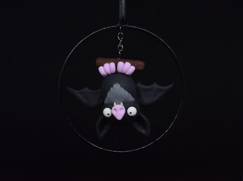 May include: A whimsical, handmade bat ornament. The bat is black with gray accents, large white eyes, and a pink nose and ears. It hangs upside down from a brown branch with pink feet, suspended within a metal ring. The background is solid black.