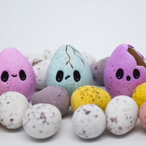 May include: A selection of speckled Easter eggs in various colours, with three cartoon-faced eggs. The eggs are pink, blue, and yellow, with black eyes and smiles. Some eggs are cracked, revealing a chocolate interior.