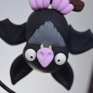 May include: A handmade, black bat figurine with large white eyes, a pink nose, and gray hair. The bat is hanging upside down, with purple feet and a brown branch. The wings are spread, and the bat is a whimsical, decorative item.