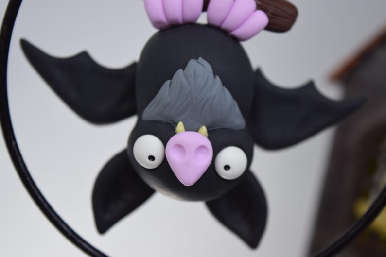 May include: A handmade, black bat figurine with large white eyes, a pink nose, and gray hair. The bat is hanging upside down, with purple feet and a brown branch. The wings are spread, and the bat is a whimsical, decorative item.
