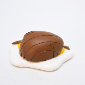 May include: A brown ceramic figurine of a bear lying on its back on a white ceramic plate shaped like a fried egg. The bear has a black nose and is detailed with lines.