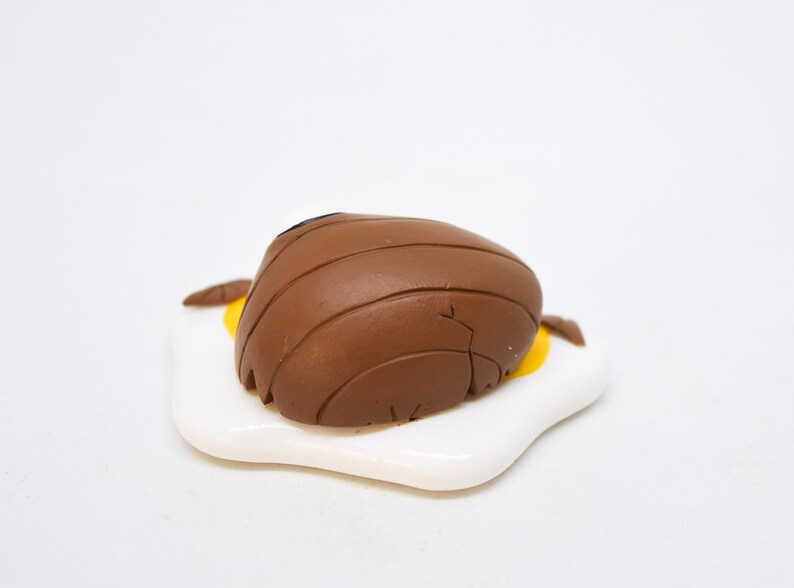 May include: A brown ceramic figurine of a bear lying on its back on a white ceramic plate shaped like a fried egg. The bear has a black nose and is detailed with lines.