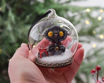 May include: A glass Christmas bauble containing a black and white monster. The monster is holding a red and white gift and a bell. The bauble is filled with white snow.