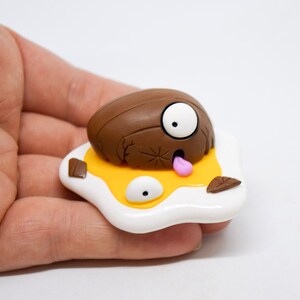 May include: A brown and white clay figurine of a cartoon character with a single large eye, a pink tongue sticking out, and a cracked shell. The character is lying on a yellow and white egg-shaped base.