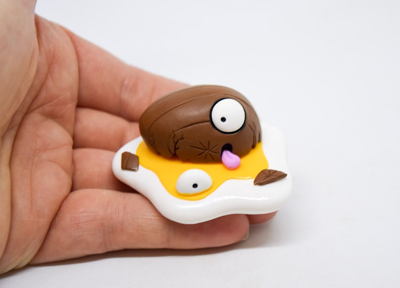 May include: A brown and white clay figurine of a cartoon character with a single large eye, a pink tongue sticking out, and a cracked shell. The character is lying on a yellow and white egg-shaped base.