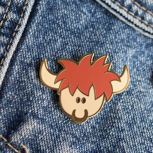 Op de afbeelding: Een gouden emaille pin met een cartoonachtige Highland koe met een rode manen en een wit gezicht. De koe heeft een vriendelijke uitdrukking met grote ogen en een glimlach.