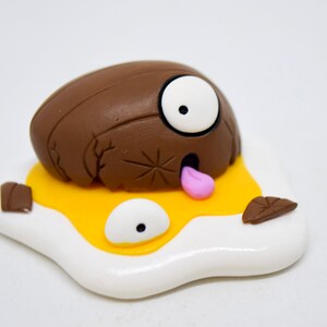 May include: A brown and white clay sculpture of a cracked egg with a googly eye and a pink tongue sticking out. The egg is lying on a white and yellow clay sculpture of a fried egg.