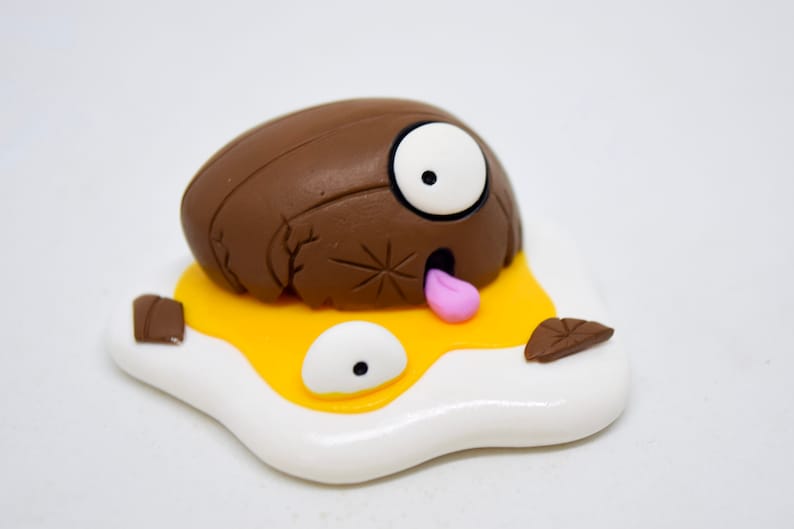 May include: A brown and white clay sculpture of a cracked egg with a googly eye and a pink tongue sticking out. The egg is lying on a white and yellow clay sculpture of a fried egg.