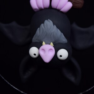 May include: A handcrafted, whimsical bat figurine. The bat is black with large white eyes, a light purple nose and ears, and gray fur. The bat is hanging upside down against a black background.