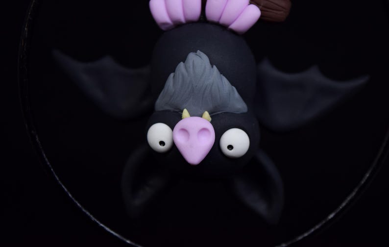 May include: A handcrafted, whimsical bat figurine. The bat is black with large white eyes, a light purple nose and ears, and gray fur. The bat is hanging upside down against a black background.