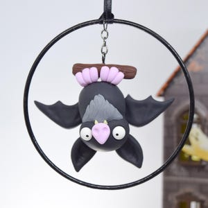May include: A handcrafted Halloween ornament featuring a black bat with gray accents, pink ears, and a pink nose, hanging upside down from a black hoop. The bat is made of clay and has large white eyes. A spooky house is in the background.