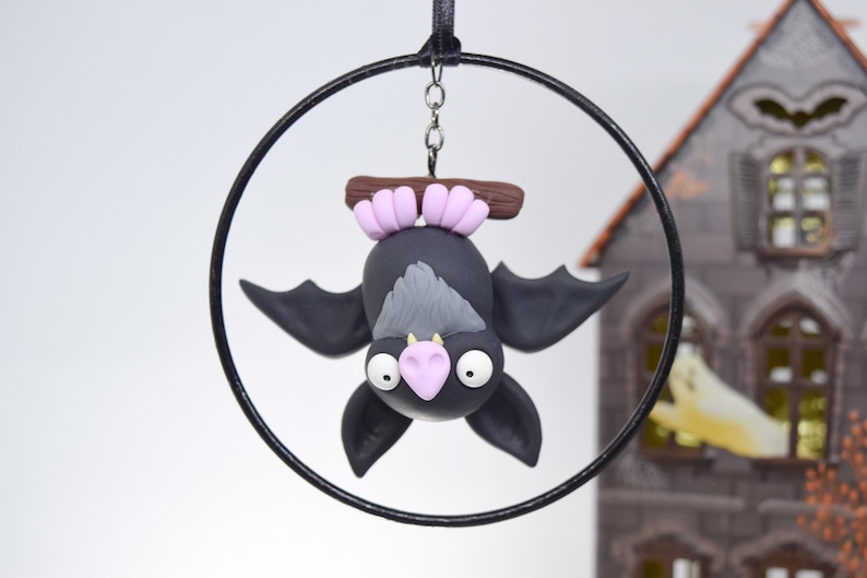 May include: A handcrafted Halloween ornament featuring a black bat with gray accents, pink ears, and a pink nose, hanging upside down from a black hoop. The bat is made of clay and has large white eyes. A spooky house is in the background.