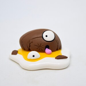 May include: A brown and white clay figurine of a cartoon character with one eye, a tongue sticking out, and a cracked shell. The character is lying on a white and yellow clay egg.