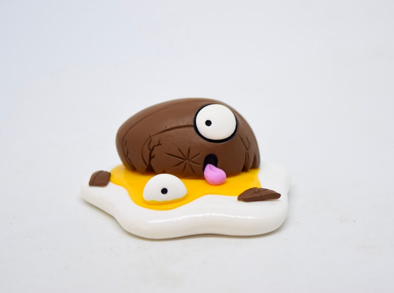 May include: A brown and white clay figurine of a cartoon character with one eye, a tongue sticking out, and a cracked shell. The character is lying on a white and yellow clay egg.