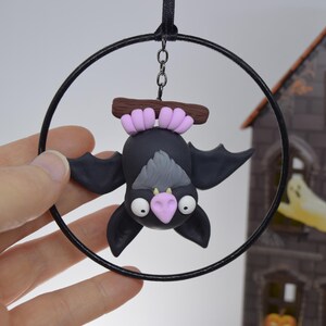 May include: A black bat ornament with a gray belly and pink nose, hanging upside down from a black hoop. The bat is attached to a brown branch with pink accents. A black ribbon and chain are used for hanging. The background includes a decorative Halloween house.