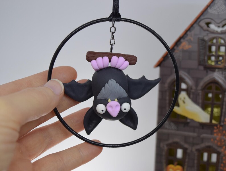 May include: A black bat ornament with a gray belly and pink nose, hanging upside down from a black hoop. The bat is attached to a brown branch with pink accents. A black ribbon and chain are used for hanging. The background includes a decorative Halloween house.
