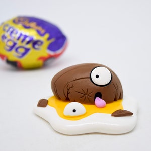 May include: A brown chocolate egg with a white eye and pink tongue, cracked open and lying on a yellow yolk with a white egg white. The egg is surrounded by brown pieces of shell.