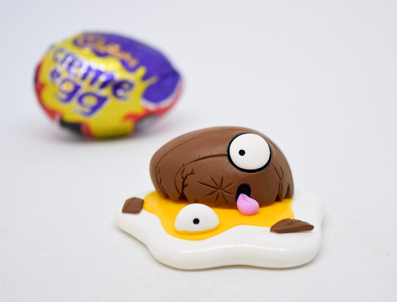 May include: A brown chocolate egg with a white eye and pink tongue, cracked open and lying on a yellow yolk with a white egg white. The egg is surrounded by brown pieces of shell.