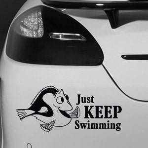 May include: Black and white vinyl car decal showing a cartoon fish and the words "Just KEEP Swimming". The sticker is on the back of a white car. The fish has a cheerful expression.
