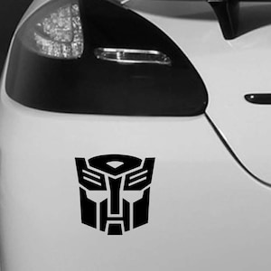 May include: Black vinyl Autobot symbol sticker, a robot face design, applied to the rear of a white car. The sticker is positioned near the taillight. The Autobot symbol is a well-known design from the Transformers series.