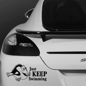 May include: A black and white vinyl decal of a cartoon fish with the words "Just KEEP Swimming" on the back of a white car. The car has a black spoiler and rear lights.