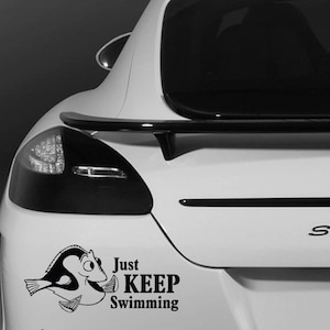 May include: A black and white vinyl decal of a cartoon fish with the words "Just KEEP Swimming" on the back of a white car. The car has a black spoiler and rear lights.