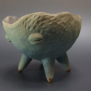 以下が含まれることがあります： 手作りの三本足のセラミックボウル。表面は質感があり、淡い青とベージュのグラデーションカラー。ボウルは、顔のような外観を持つユニークで様式化されたデザインです。足の先端は金色です。