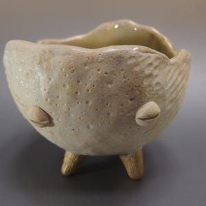以下が含まれることがあります： 手作りのオフホワイトのセラミックボウル。表面はテクスチャ加工が施され、3つの小さな金色の脚が付いています。ボウルは独特の有機的な形状で、側面に2つの装飾的な丸みを帯びた突起があります。