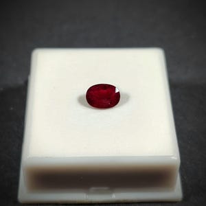 Può includere: Un'intensa gemma di rubino ovale, di colore rosso scuro, presentata su una base rettangolare bianca. La gemma è al centro, su uno sfondo scuro. Il rubino mostra il suo colore intenso e la sua chiarezza, catturando la luce.
