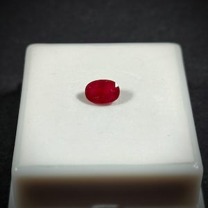 May include: A vibrant, oval-cut ruby gemstone, displayed on a white surface. The ruby exhibits a deep red hue, with a polished surface. The gemstone is set against a dark background, highlighting its color and clarity. A precious stone.