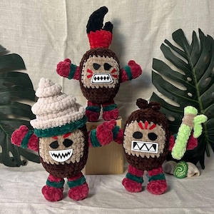 May include: Three crocheted plush toys with brown bodies, red arms and legs, and different facial expressions. The toys are wearing green and white hats. One toy is holding a green stick.