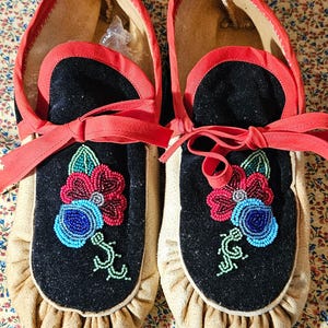 May include: A pair of tan suede moccasins featuring black velvet vamps adorned with intricate floral beadwork in red, blue, and green. The shoes include red fabric trim along the collar and matching red ribbon laces tied at the front.