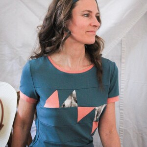 May include: A teal blue t-shirt with a pink and white geometric design. The shirt has a pink trim around the neckline and sleeves.