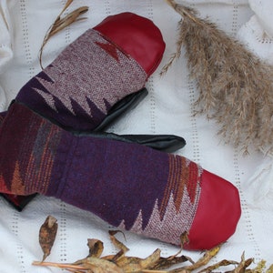 Peut inclure: Une paire de mitaines à motif rouge et violet avec des paumes en cuir. Les mitaines sont pliées ensemble et posées sur un fond de tissu blanc.