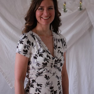 May include: A woman wearing a white dress with a black floral pattern. The dress has a V-neckline and short sleeves.