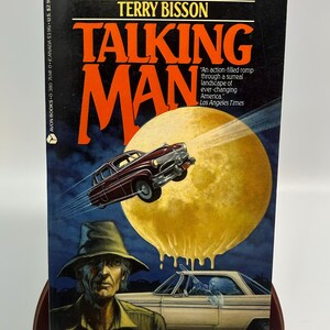 May include: A paperback book titled "Talking Man" by Terry Bisson. The cover art features a vintage car, a man in a hat, and a large yellow moon. The book's title is in large orange letters.