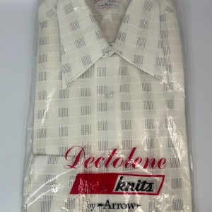May include: Cream-colored button-down shirt with a grid pattern of dark gray lines, packaged in a clear plastic bag. The shirt has a pointed collar and long sleeves. The packaging includes the text "Dectolene knits by Arrow".