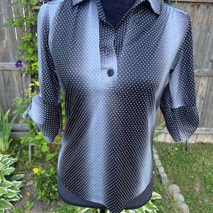 May include: A black and gray polka dot blouse with a collared neckline and three-quarter sleeves. The blouse has a button-down front and is displayed on a mannequin. The background includes a wooden fence and greenery.