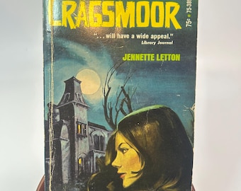 May include: A vintage paperback book titled "Cragsmoor" by Jennette Letton. The cover features a woman's profile, a spooky mansion, and a full moon against a dark blue and green background. The book is displayed on a wooden stand.