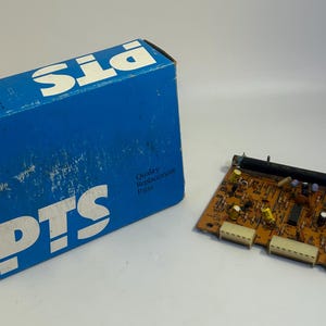 May include: A blue box with the text "PTS" printed on it. The box is sitting next to a small circuit board with electronic components. The circuit board is brown and has a black wire attached to it. The text "Quality Replacement Parts" is printed on the box.