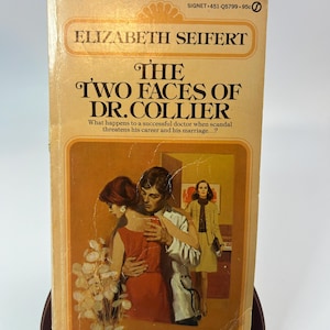 May include: Vintage paperback book titled "The Two Faces of Dr. Collier" by Elizabeth Seifert. The cover features a couple embracing, with a woman in a red dress and a man in a white coat. The book is displayed on a wooden stand.