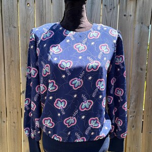 May include: A long-sleeved blouse in a deep navy blue featuring a repeating floral pattern in shades of magenta, light blue, and white. The garment has a round neckline, gathered shoulders, and a solid navy blue ribbed hem and sleeve cuffs.
