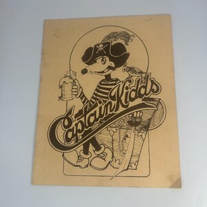 May include: A vintage print featuring a cartoon pirate mouse holding a mug of beer. The mouse wears a pirate hat and striped shirt. The text "Captain Kidds" is prominently displayed in a stylized font, with a treasure chest graphic.