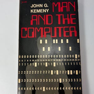 May include: A vintage book titled "Man and the Computer" by John G. Kemeny. The cover is black with red lettering and a binary code design. The price tag shows $1.45.