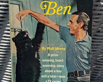 May include: Vintage paperback book cover for "Gentle Ben" by Walt Morey. The cover features a man interacting with a black bear. The title is in yellow, with the author's name and a description of the story. The book is a Starline Edition.