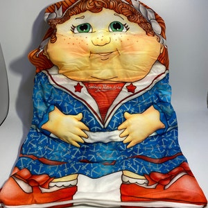 May include: A Cabbage Patch Kids pillow featuring a red-haired girl wearing a blue sailor outfit with anchor print and red shoes. The pillow has a zipper and the text "Cabbage Patch Kids" is printed on the front.