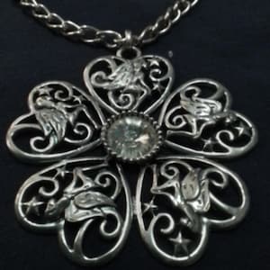 May include: Silver-toned pendant necklace featuring a floral design with heart-shaped petals. Each petal has a detailed, stylized bird and star accents. A clear, round gem is set in the center. The necklace has a chain.