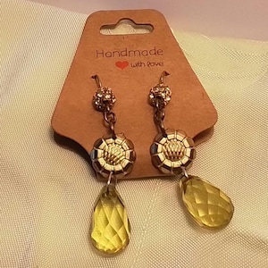 May include: A pair of handmade earrings with a brown tag that reads "Handmade with love". The earrings have gold-colored accents, a faceted yellow teardrop-shaped gem, and a decorative flower-shaped center.