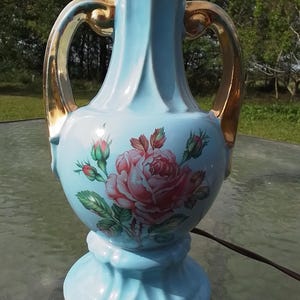 May include: A vintage ceramic table lamp in a light blue hue, featuring a large pink rose floral design. The lamp has gold-colored handles and accents, with a bulb fixture at the top. The lamp is a decorative lighting fixture.