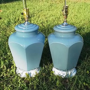May include: Two matching blue-green table lamps with a geometric design. The lamps have a rounded top and a white base. The lamps are sitting on green grass. The lamps have a brass-colored lamp holder.